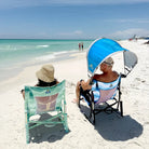 A man sitting in a beach rocker on the beach with the saybrook blue sunshade accessory attached to the chair. 