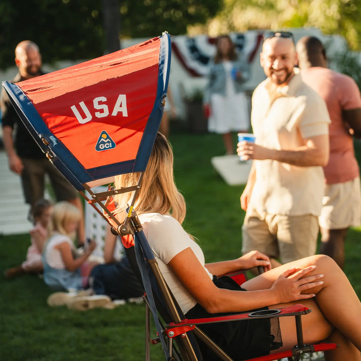 A woman sitting in a Comfort Pro Rocker chair with a USA Sunshade Accessory attached at a backyard party. 