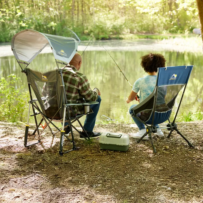 Two people fishing at a wooded lakeside, seated in a SunShade Rocker with canopy and a ComPack Rocker with mesh back.