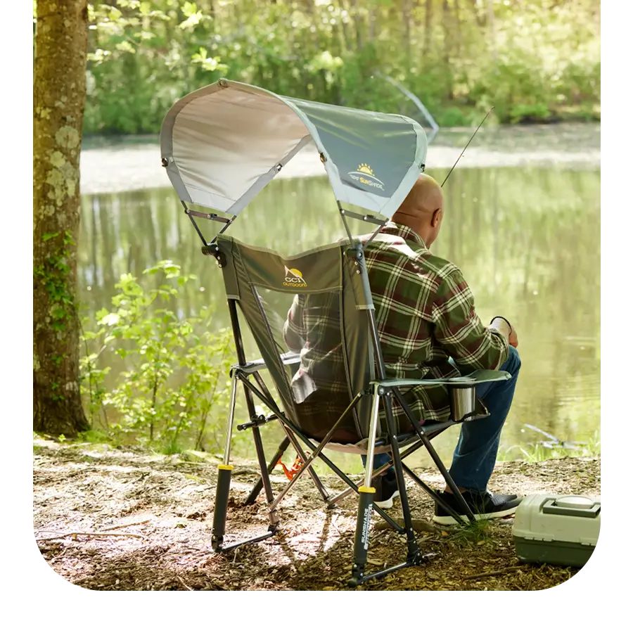 Man fishing by a calm lake, seated in a Sunshade Rocker with a gray canopy for sun protection.