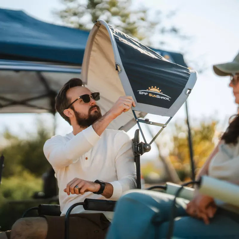 A man wearing sunglasses adjusts a dark gray SPF Sunshade attached to his chair, with another person seated nearby under a canopy.