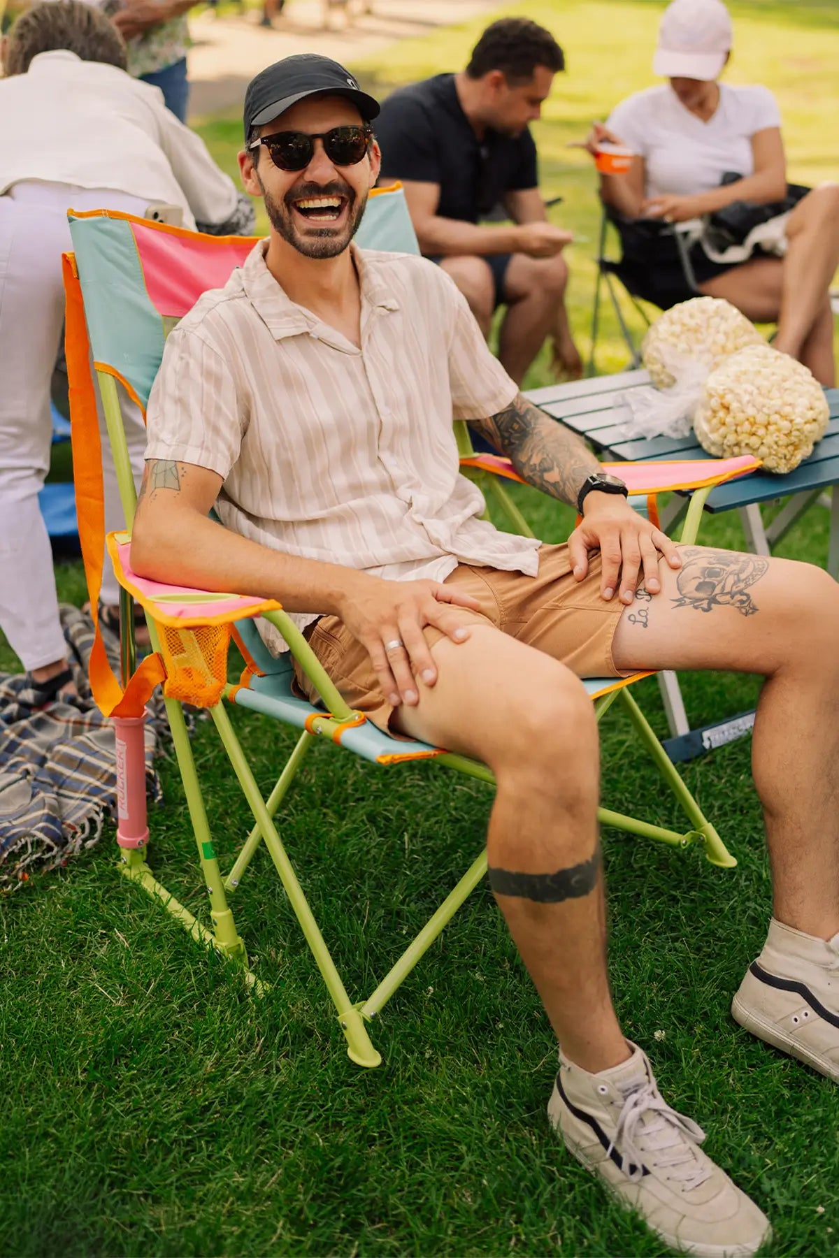 A man sitting outdoors in a colorful folding camping chair on grass, wearing a light beige striped short-sleeve button-up shirt, tan shorts, a black cap, and sunglasses. 