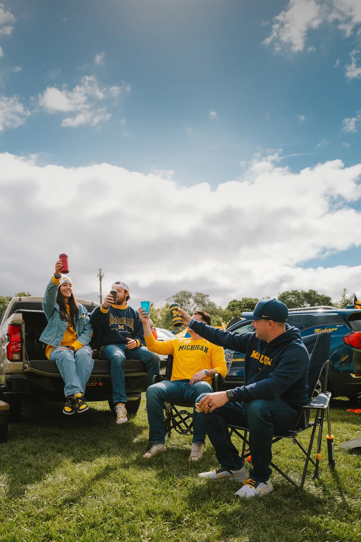 A group of friends relax in a grassy outdoor setting, sitting on the tailgate of a pickup truck and in folding chairs under a blue sky with clouds.
