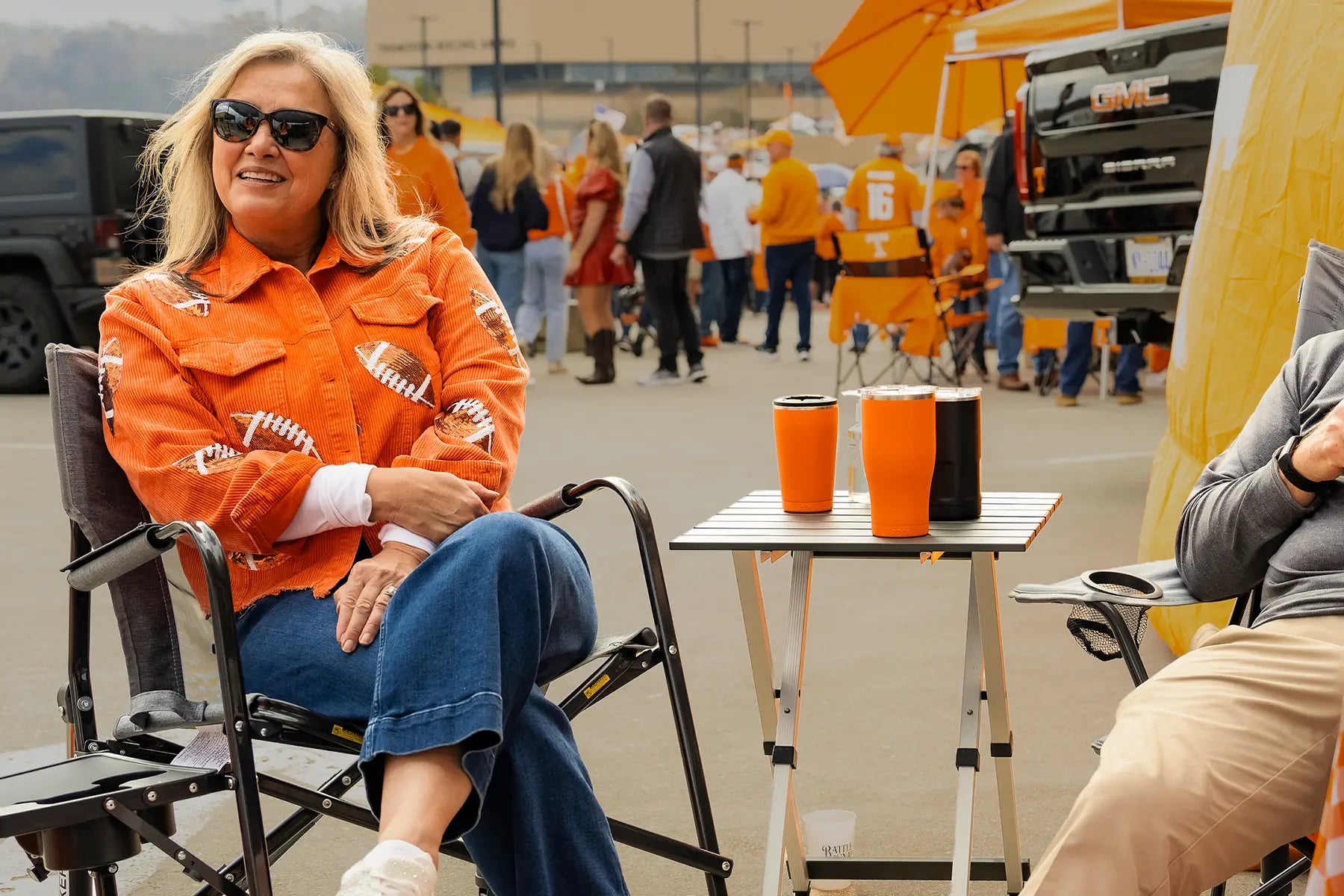A woman sitting in a freestyle rocker with side table at a parking lot tailgate. 