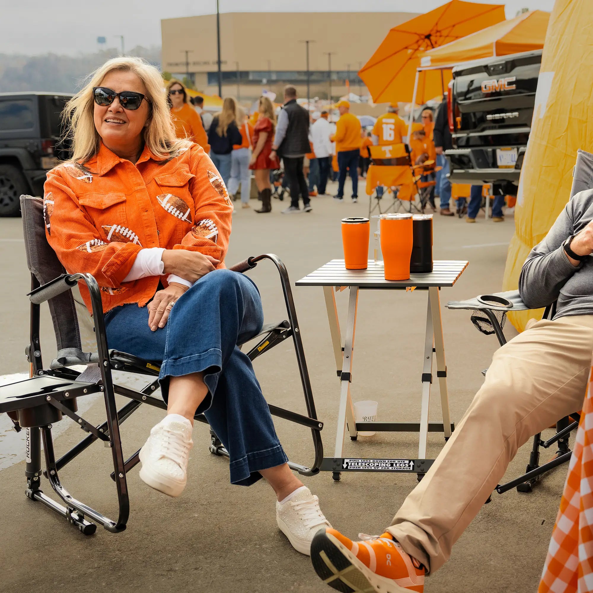 A woman sitting in a freestyle rocker with side table at a parking lot tailgate. 