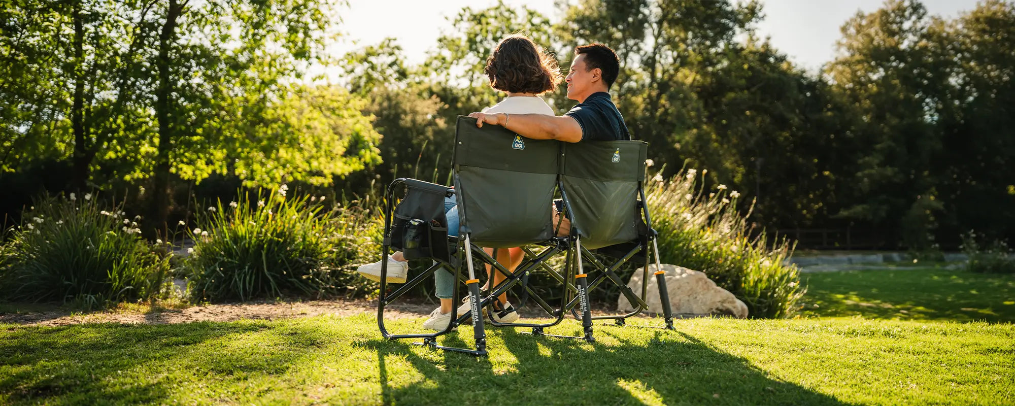 A man and woman sitting on a Tandem Rocker together at a park. 