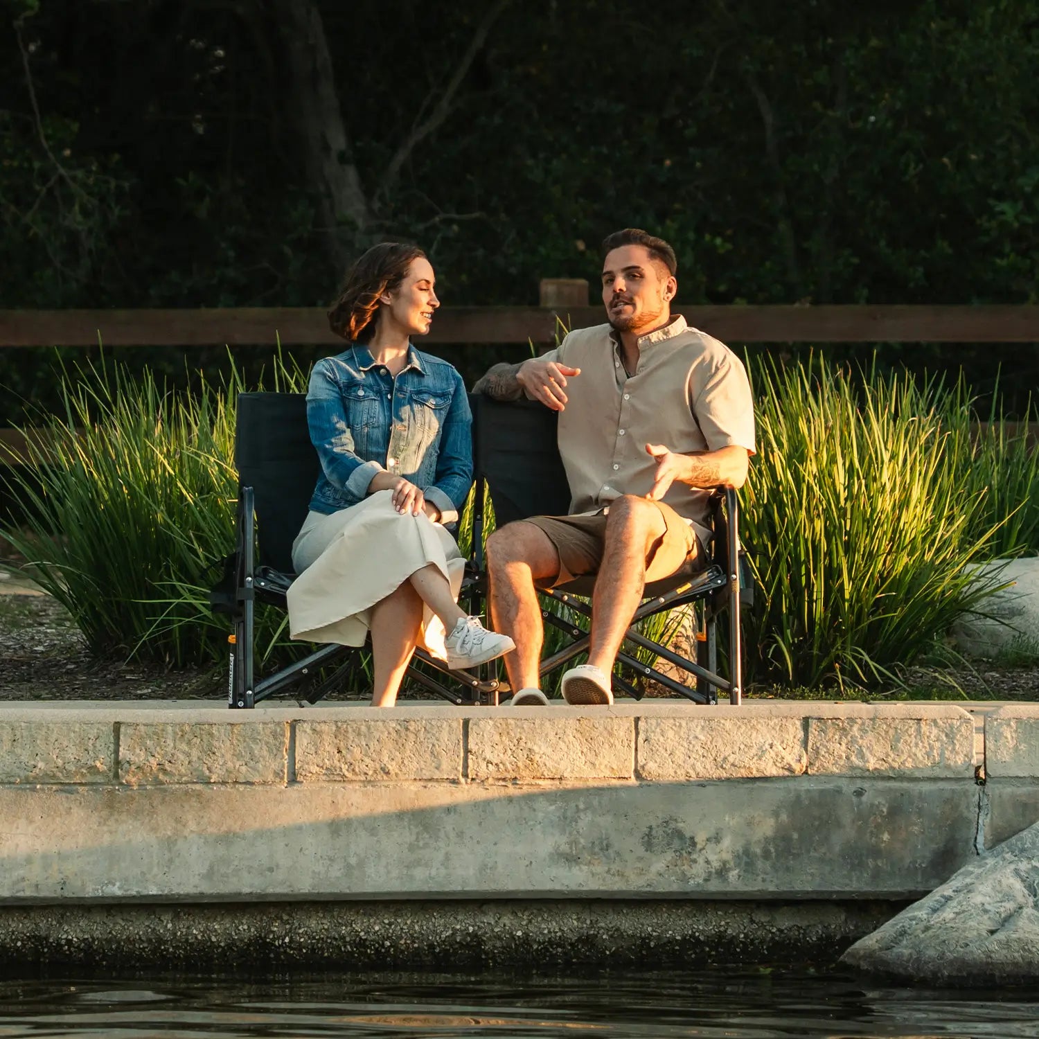 A couple sitting together and talking as they sit on a Tandem Rocker near a pond. 