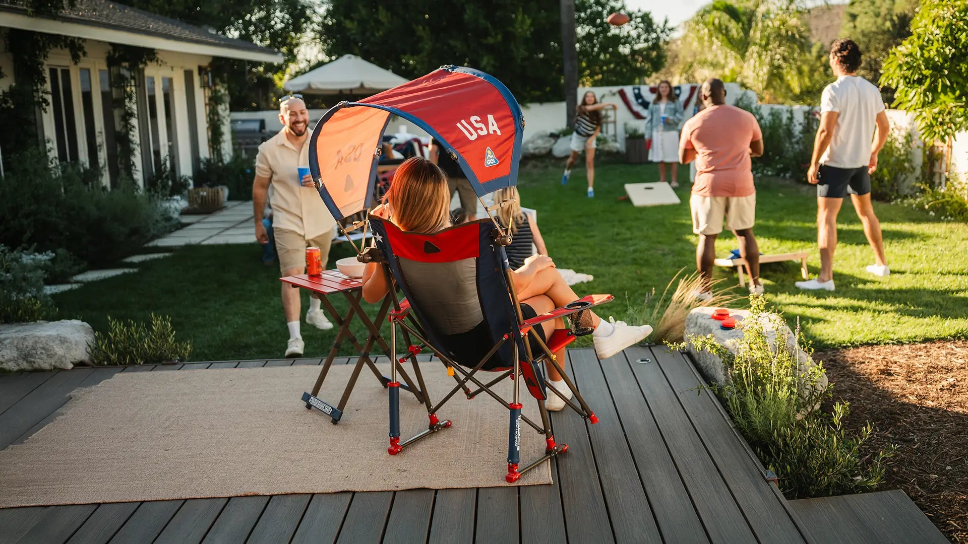 A woman sitting in a USA Comfort Pro Rocker while watching a group play football at a party. 