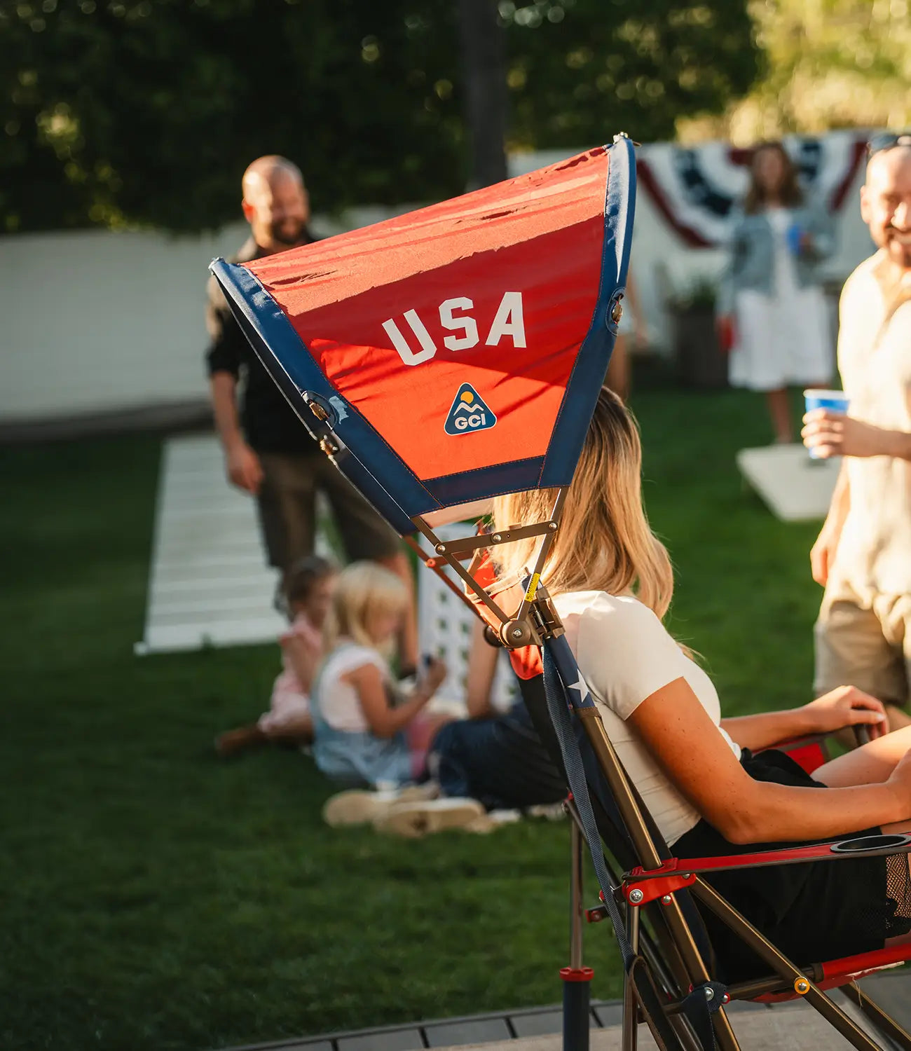 A woman attending a backyard party while sitting in a USA themed Comfort Pro Rocker with a USA Sunshade Accessory attached. 