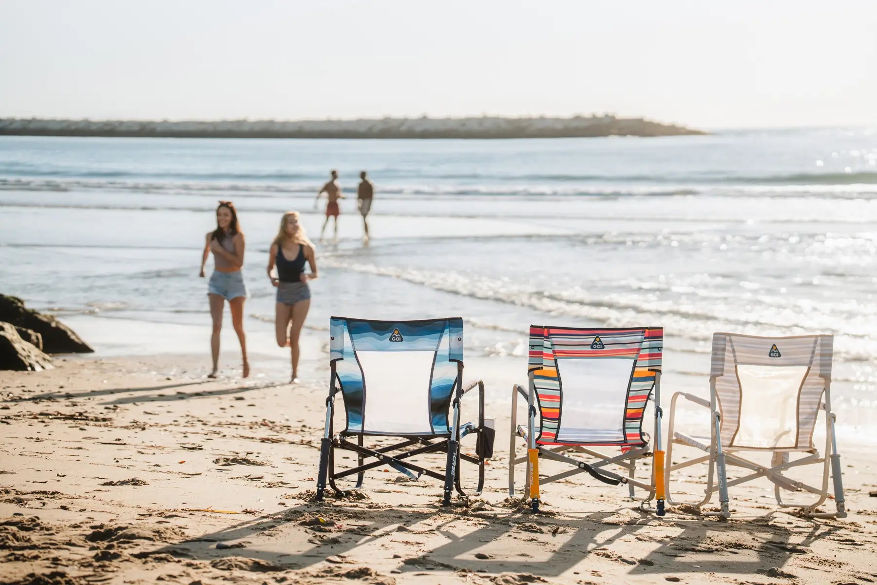 Three beach rocker chairs on a sandy beach, with people walking and playing in the ocean behind them.