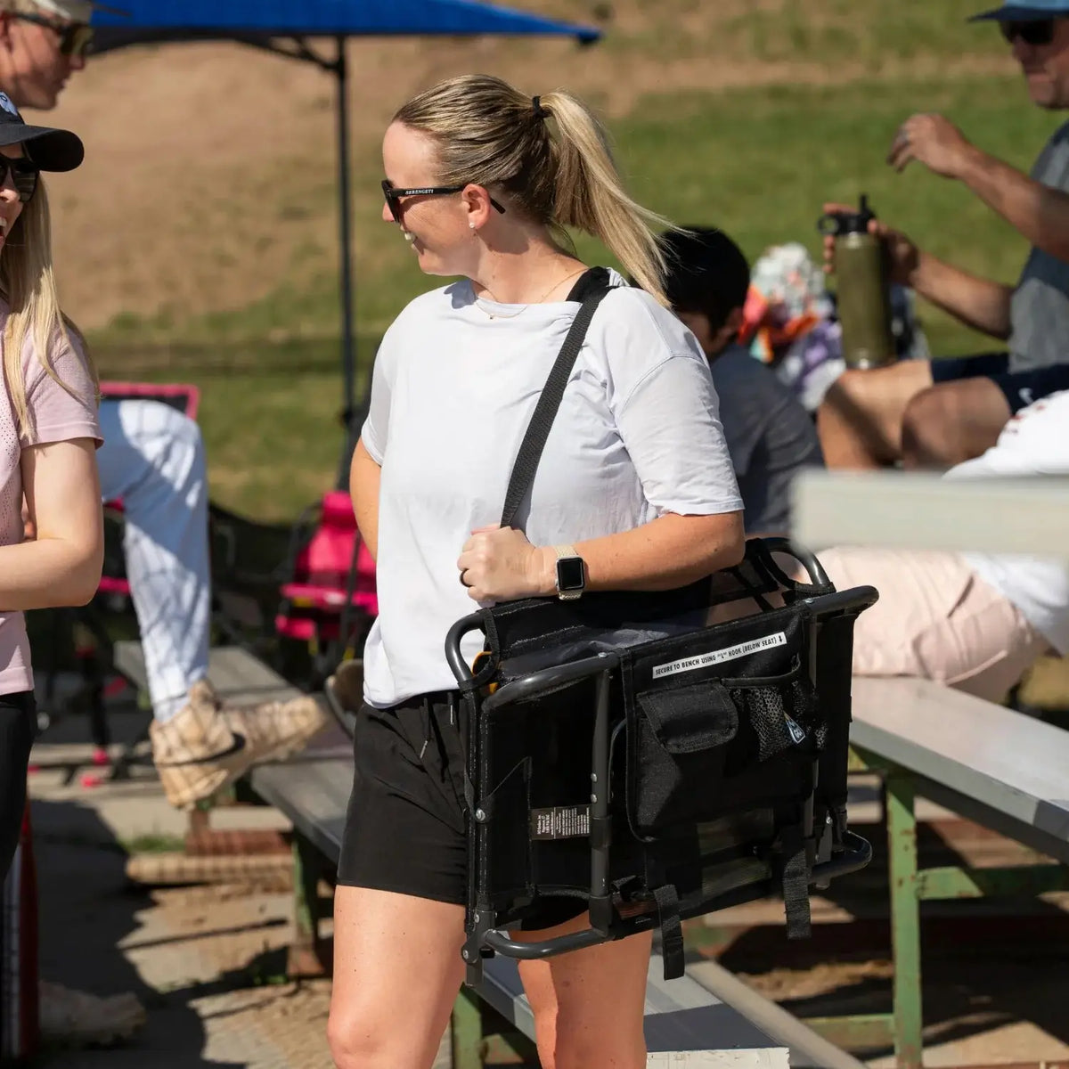 Woman carrying a folded Big Comfort Stadium Chair 2.0 over her shoulder using the built-in strap while walking through bleacher seating.