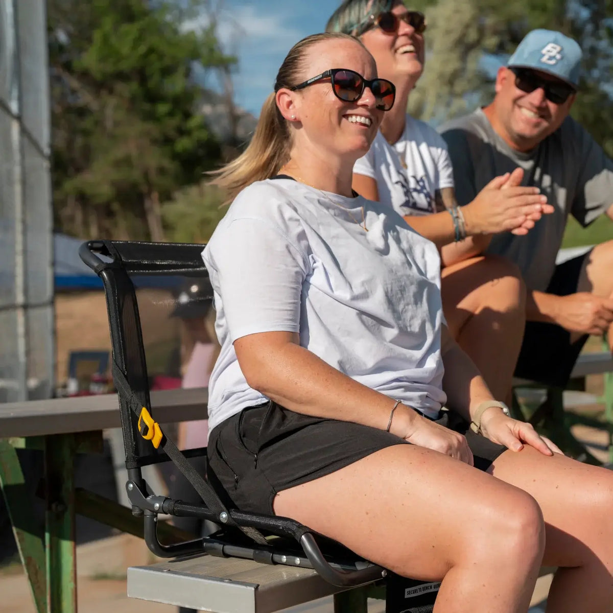 Woman sits in a Big Comfort Stadium Chair 2.0 secured to a bleacher bench, smiling alongside others.