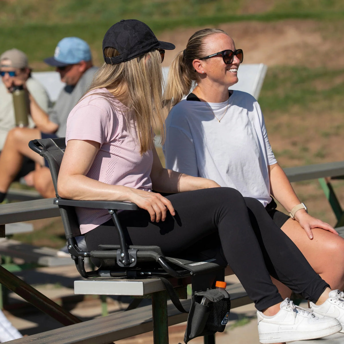 Woman sits in a dark charcoal Big Comfort Stadium Chair with Armrests 2.0 on bleachers, smiling alongside another person.