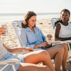 Woman seated in a GCI Big Surf with Slide Table in light blue mosaic, eating a sandwich alongside two friends on the beach.