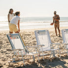 A GCI Big Surf chairs in retro beach stripes sits alongside two light blue mosaic chairs, with people playing football on the beach behind them.