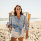 Woman walks along the beach with a GCI Big Surf with Slide Table in retro beach stripes worn backpack-style, holding a canned drink.