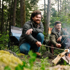 Two campers roast marshmallows over a campfire in the woods, one seated in an indigo Comfort Pro Insulated Rocker.