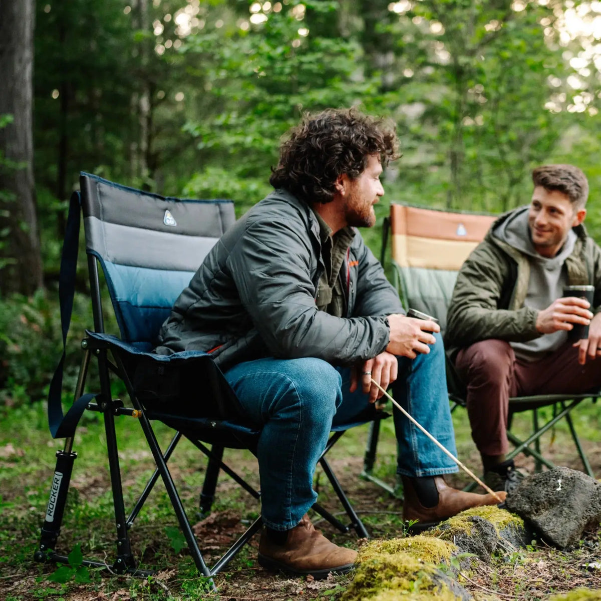Man sits in an indigo Comfort Pro Insulated Rocker holding a metal cup, seated beside another camper in the woods.