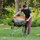 Man sets up an olive Comfort Pro Insulated Rocker with striped padding on a grassy lawn surrounded by trees.