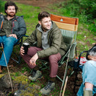 Man sits in an olive Comfort Pro Insulated Rocker holding a metal cup, surrounded by others in camp chairs outdoors.