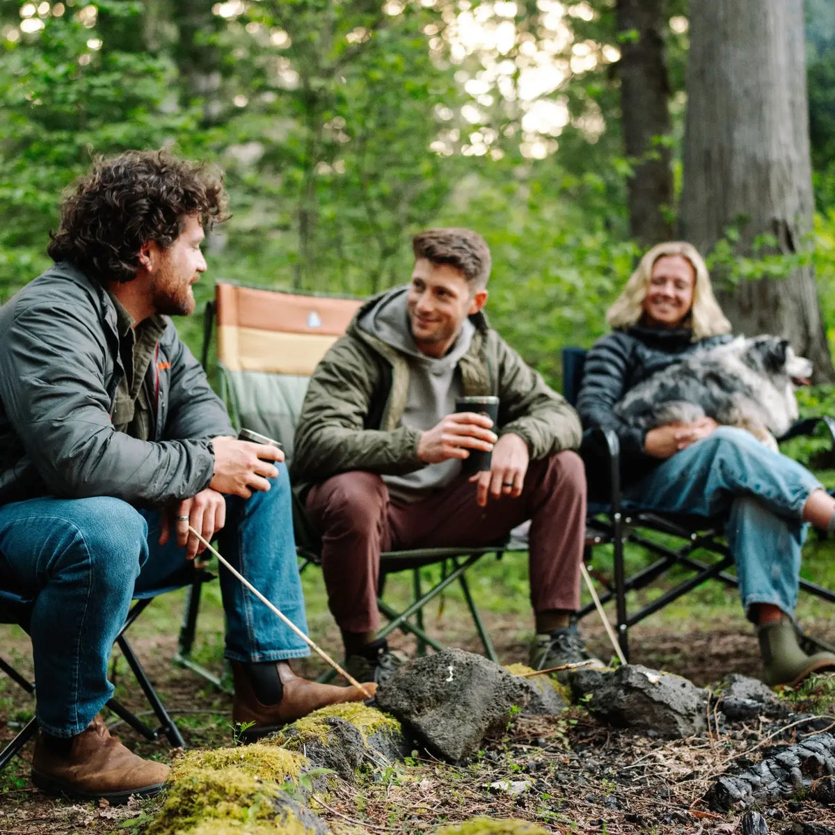Three campers sit around a fire pit in camp chairs, including an olive Comfort Pro Insulated Rocker, with a dog resting on one person's lap.