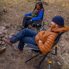 Two people sitting near a stone fire pit in an outdoor setting; one relaxes in a black freestyle rocker while the other sits in a different camping chair.