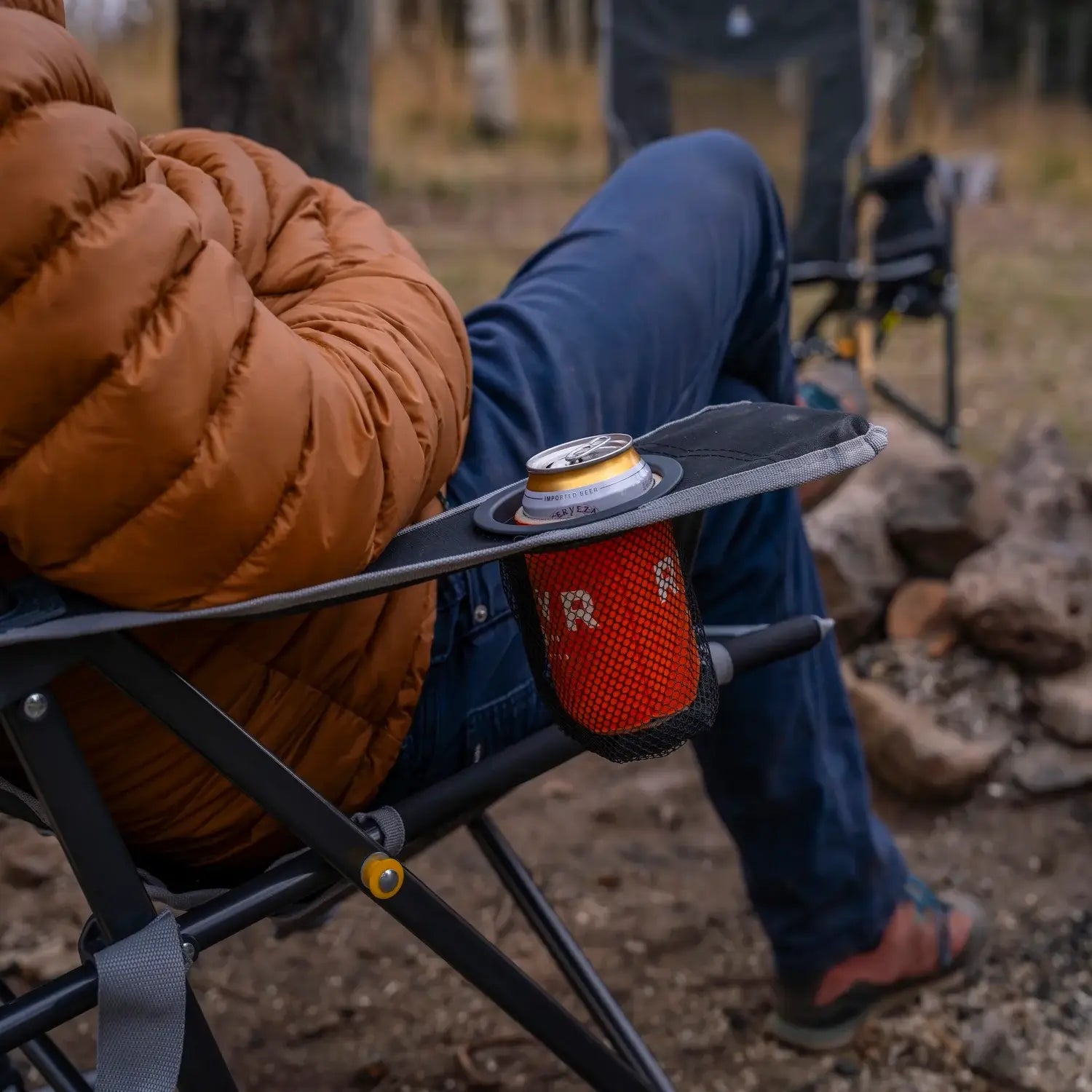 Close-up of a person reclining in a black freestyle rocker, with a beverage can resting in the chair’s mesh cup holder near a stone fire pit.