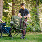 Man sets up a heathered olive Comfort Pro Rocker XL on grass near a wagon cart.