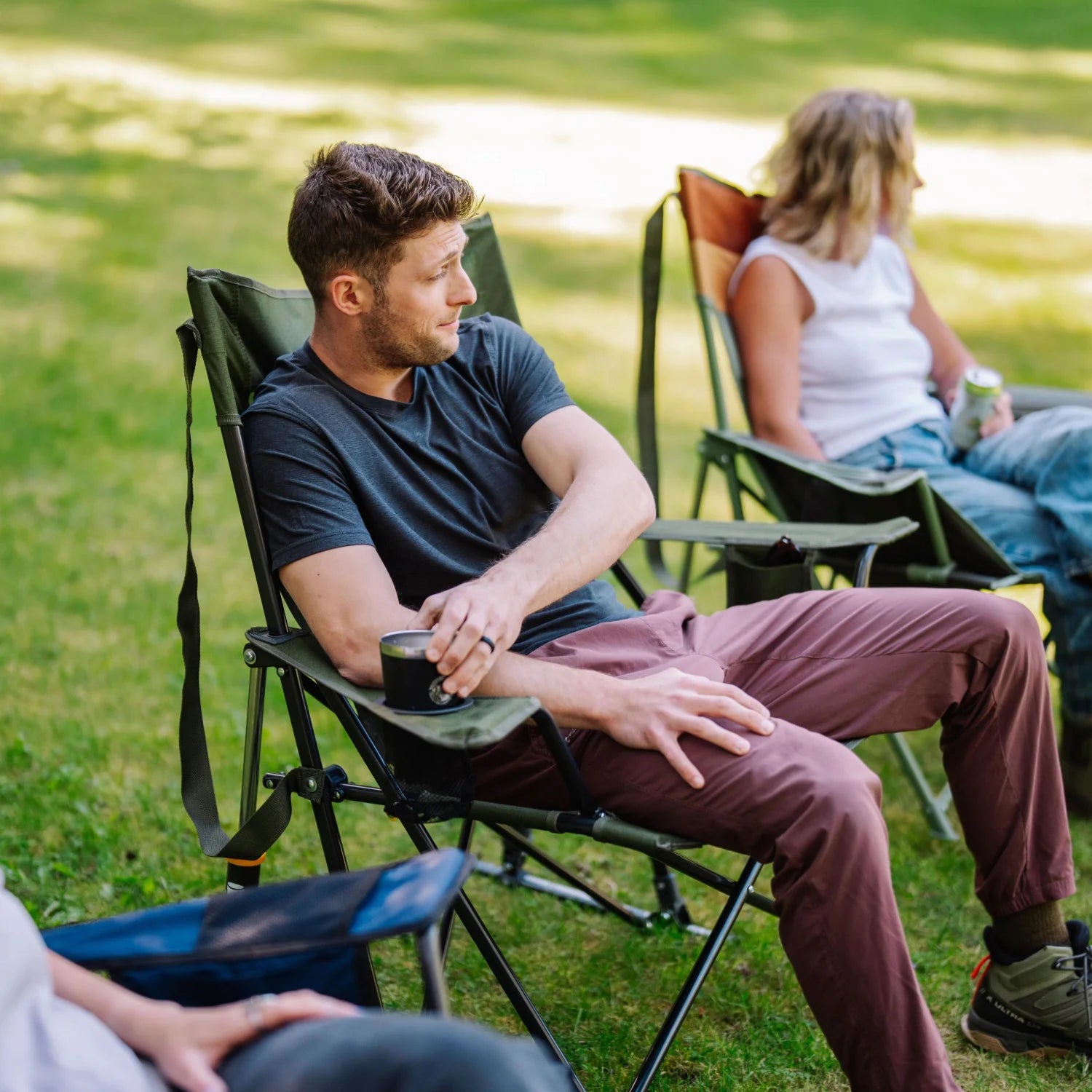 Man relaxes in a heathered olive Comfort Pro Rocker XL, holding a metal cup, with others seated in camp chairs nearby.