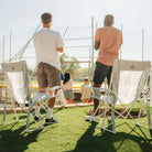 Two men standing to cheer at a softball game beside GCI Outdoor Comfort Pro Rocker XL chairs in Heathered Light Blue, rear view.