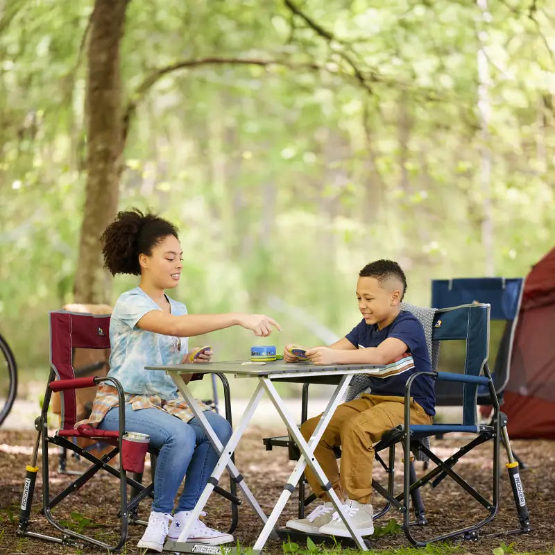 A girl and boy sit in outdoor chairs, laughing and pointing as they play, surrounded by trees and tents.