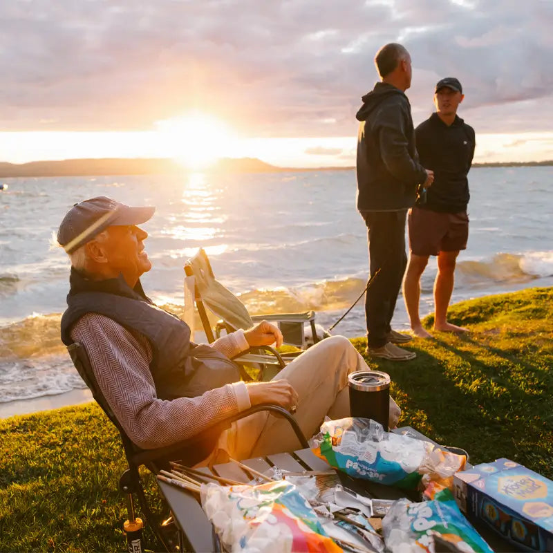 The table holds marshmallows and popcorn bags while the man enjoys the view as others fish in the background.