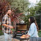A couple chats while grilling burgers on a large black grill resting on the sturdy, dark tabletop of the Compact Camp Table 25.