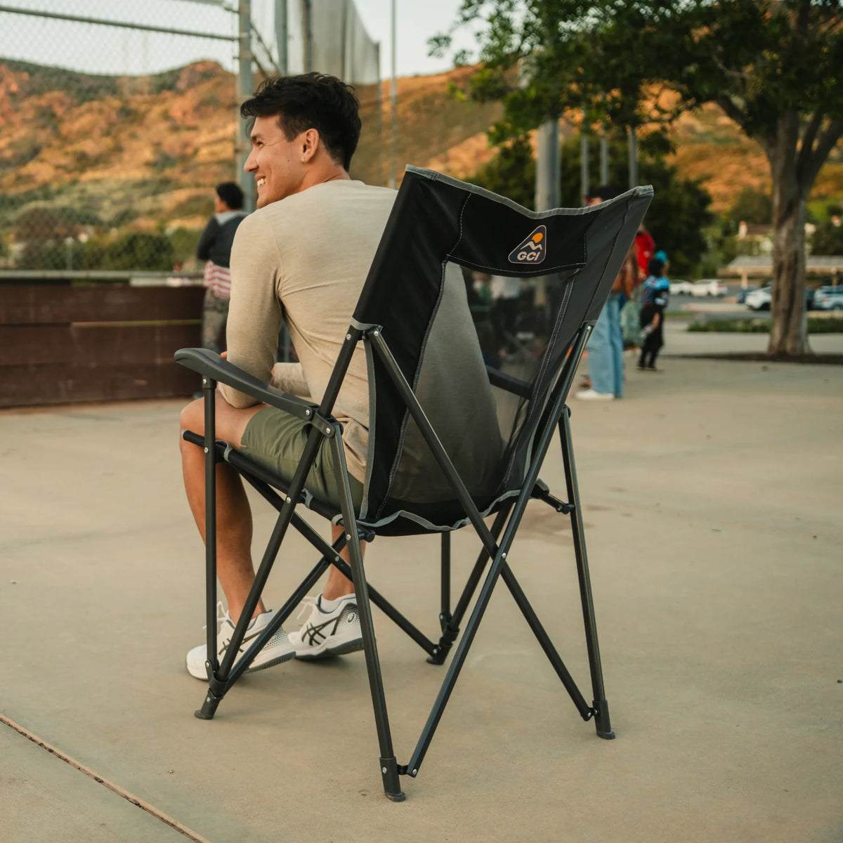 Smiling man sits in a black GCI Eazy Chair XL with a mesh back panel at an outdoor sports court.