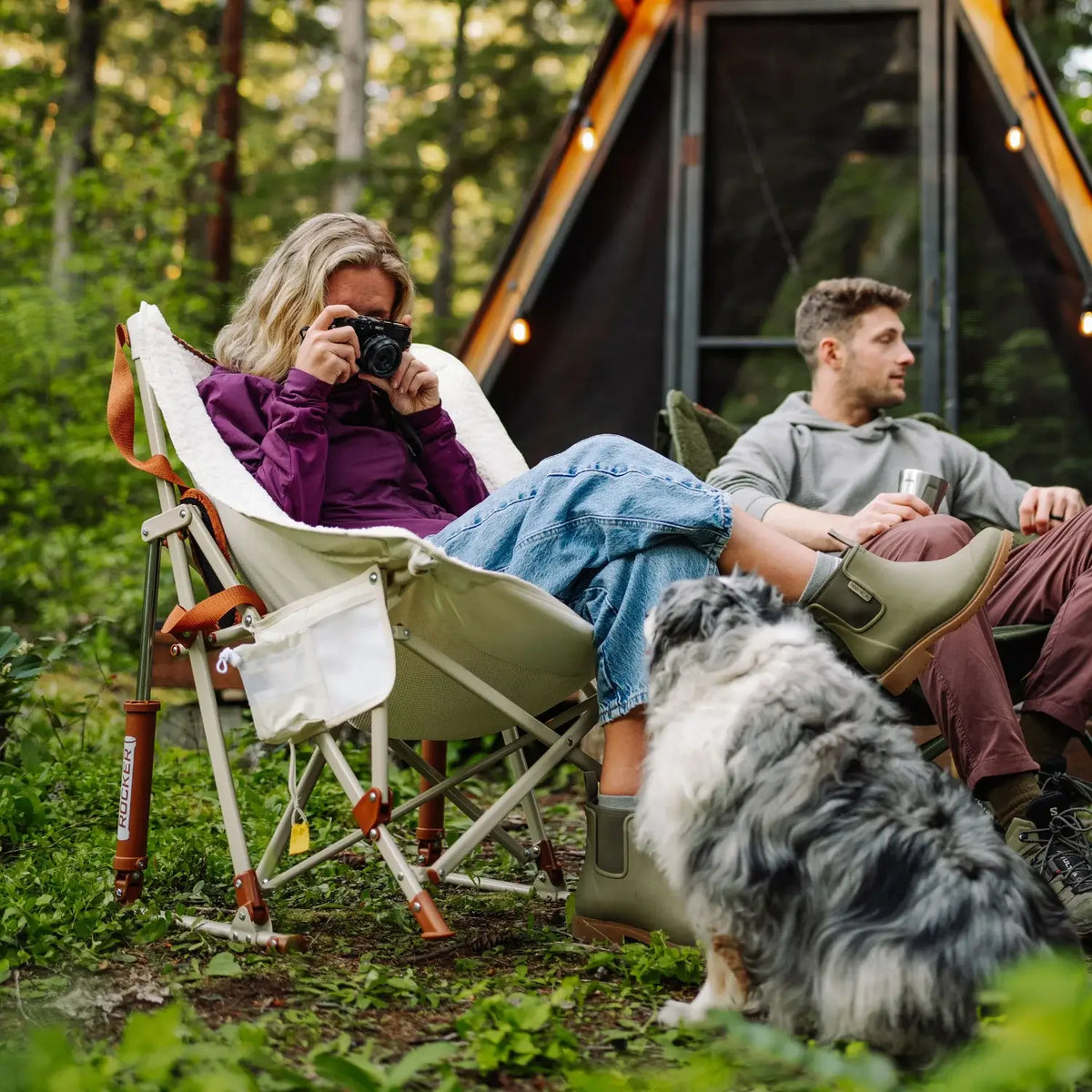 Woman photographs a dog while relaxing in a natural Fleece Rocker with her feet up, near a cabin in the woods.