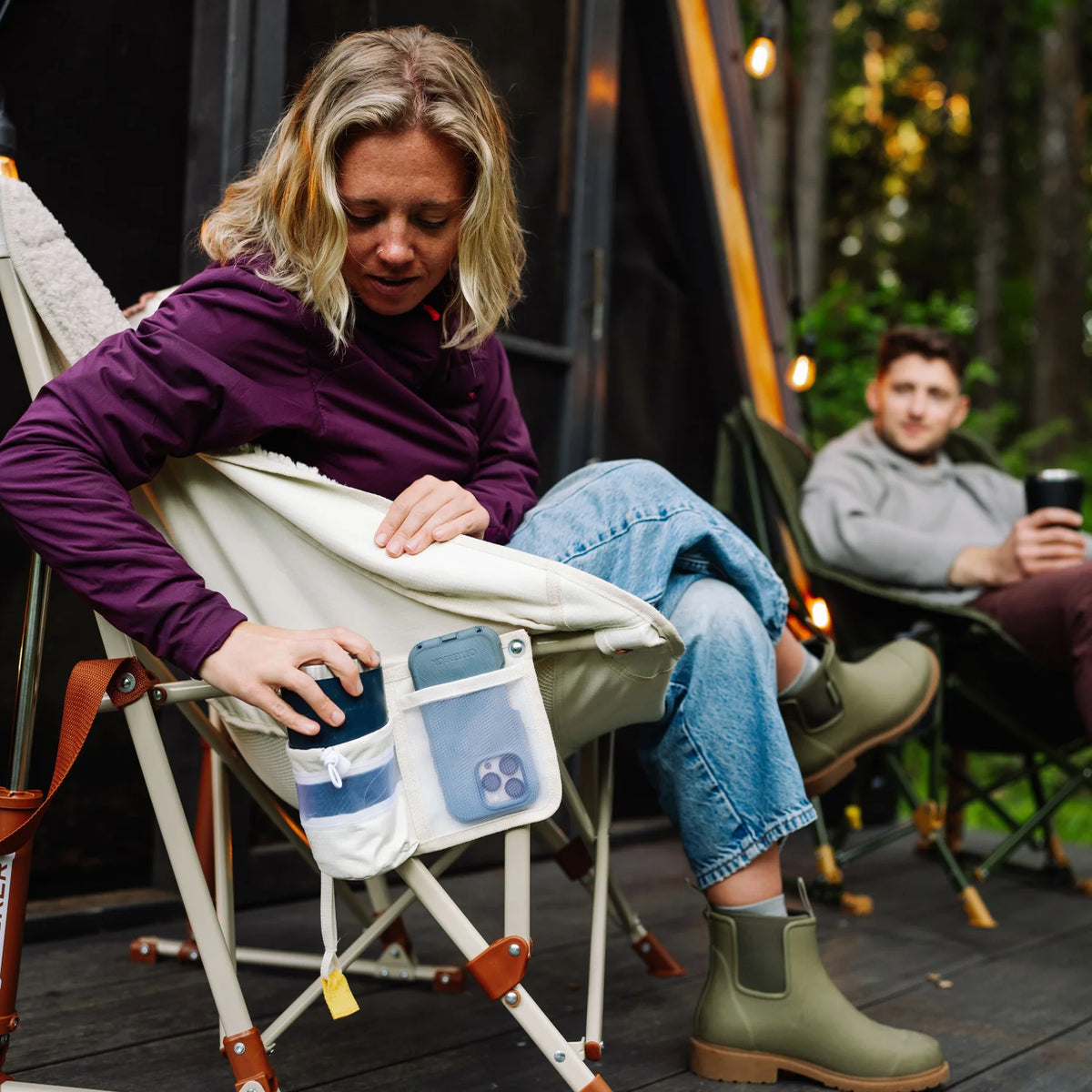 Woman reaches into the side pocket of a natural Fleece Rocker, pulling out a phone, while seated on a deck outside a cabin.
