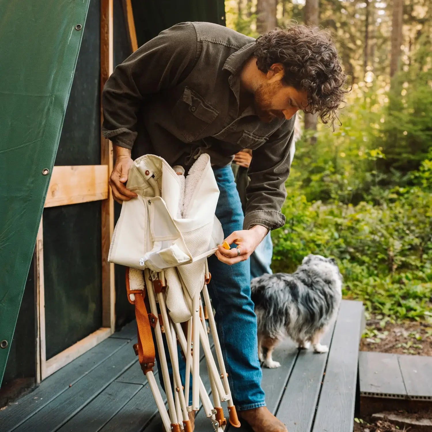 Man folds up a natural Fleece Rocker on a wooden deck, with a dog standing beside him outside a cabin.