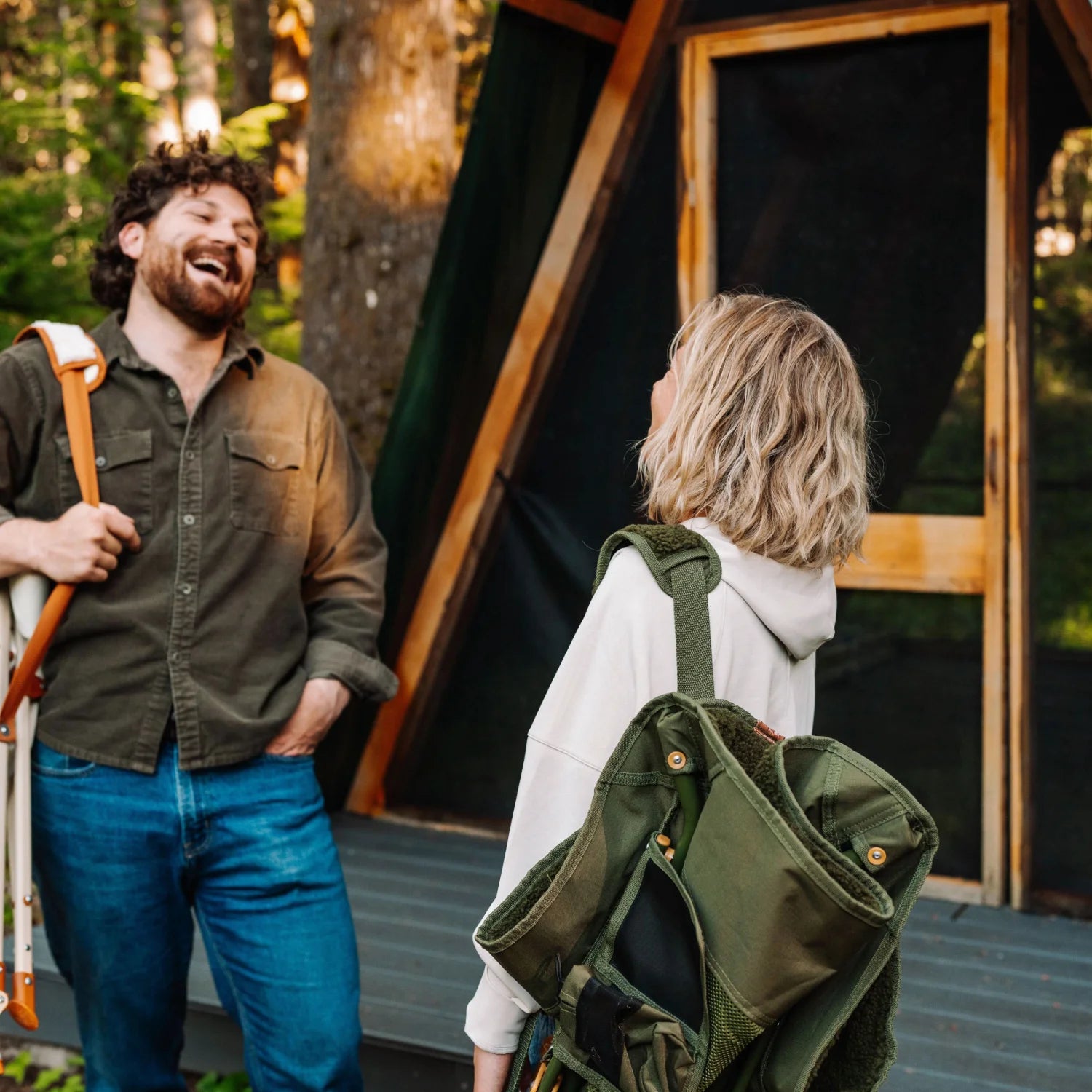 Woman carries a folded olive Fleece Rocker over her shoulder while talking with a man outside a cabin.