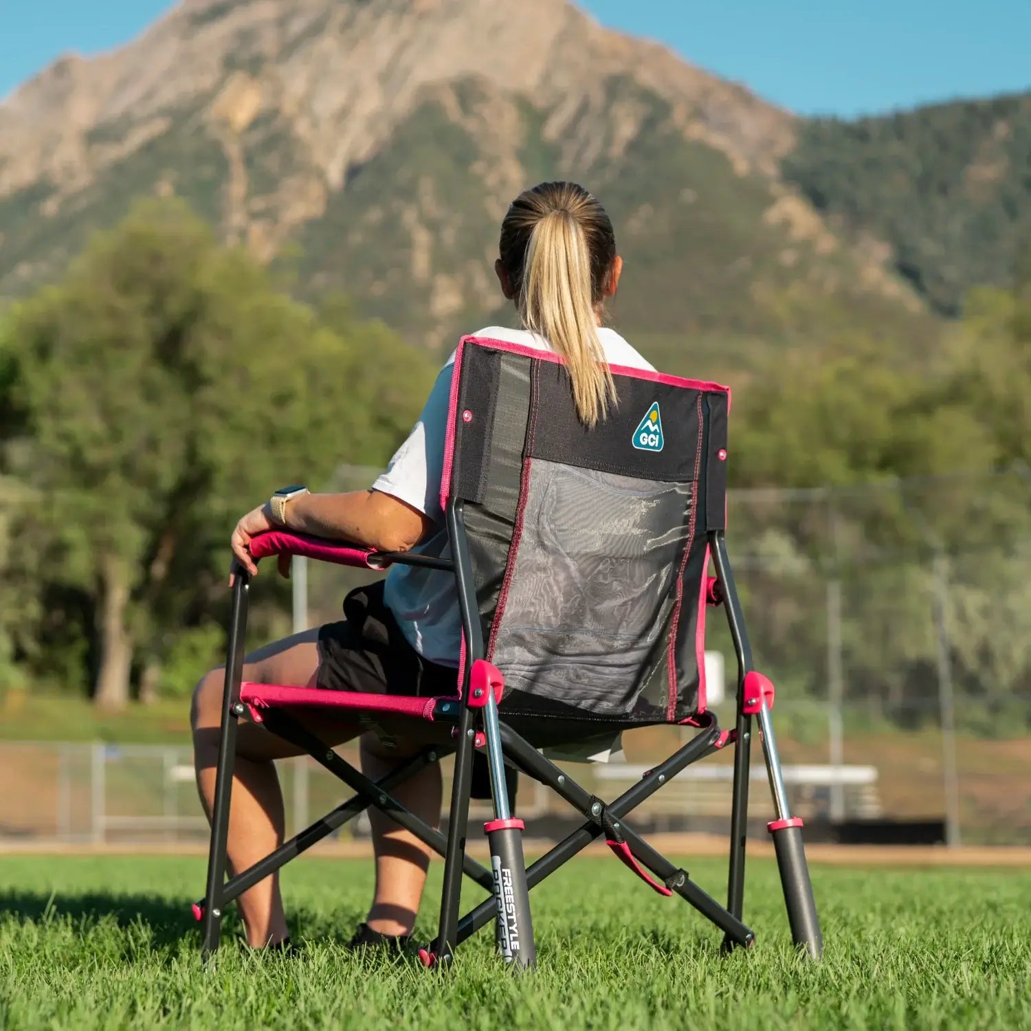 Back view of a woman seated in a pink shockwave Freestyle Rocker on a grass field, with mountains in the background.