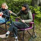 Woman roasts a marshmallow while seated in a berry Freestyle Rocker XL with Side Table, surrounded by others in camp chairs in a wooded campsite.