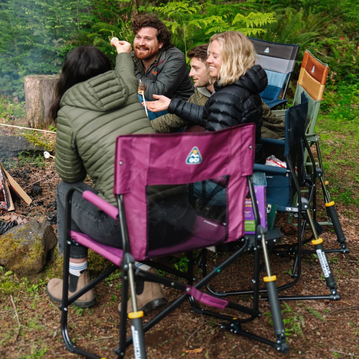 Group of friends sitting on camp chairs, including a berry Freestyle Rocker XL with Side Table, roasting marshmallows over a campfire.