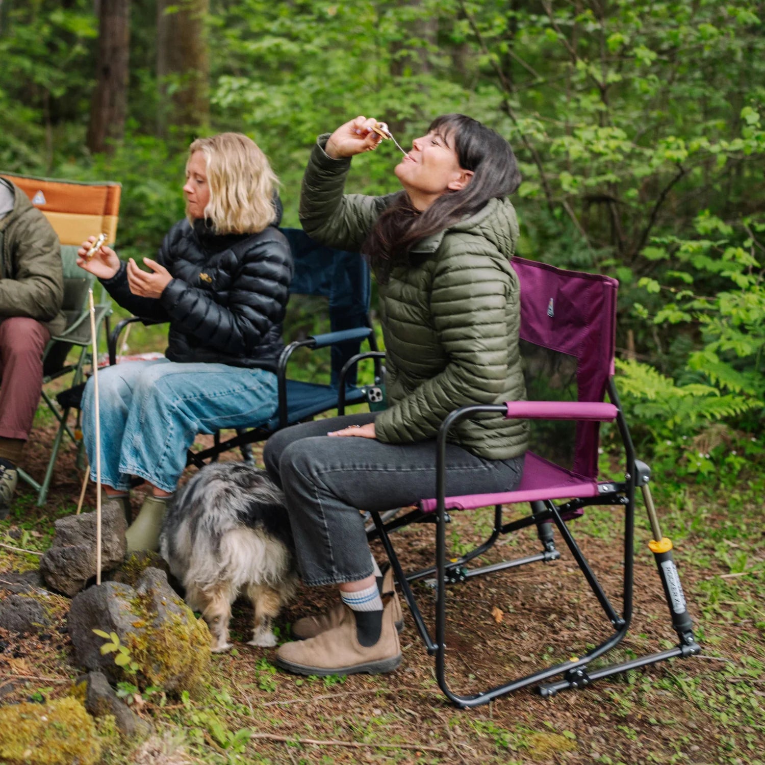 Woman eating a marshmallow while seated in a berry Freestyle Rocker XL with Side Table, with a dog and fellow campers nearby.