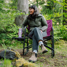 Woman relaxes in a berry Freestyle Rocker XL with Side Table in the woods, resting a snack box on the attached table.