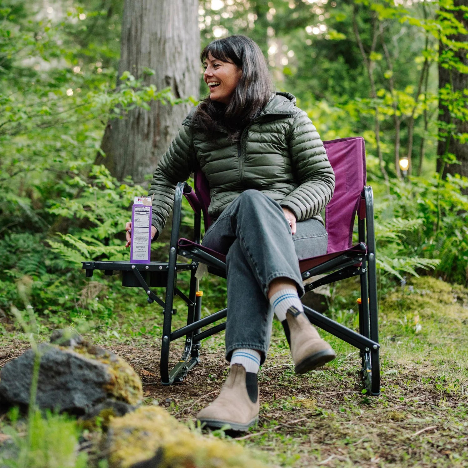 Woman relaxes in a berry Freestyle Rocker XL with Side Table in the woods, resting a snack box on the attached table.