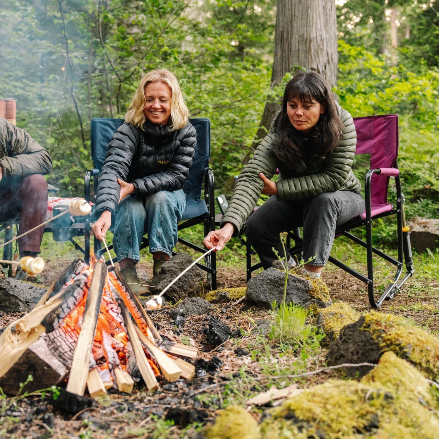 Two women roast marshmallows over a campfire, one seated in a berry Freestyle Rocker XL with Side Table.