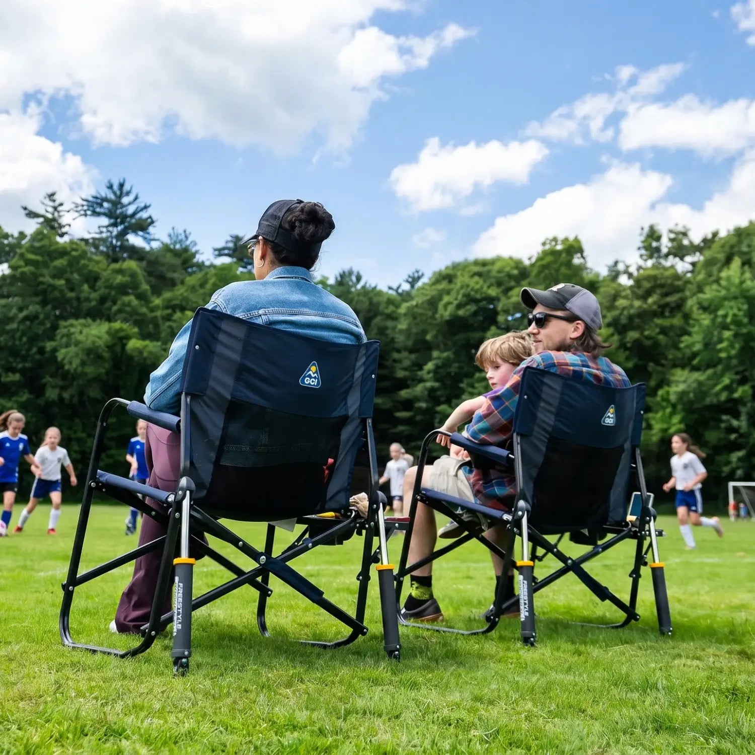 Back view of two adults and a child seated in indigo Freestyle Rocker XL chairs with Side Table on a grass field.