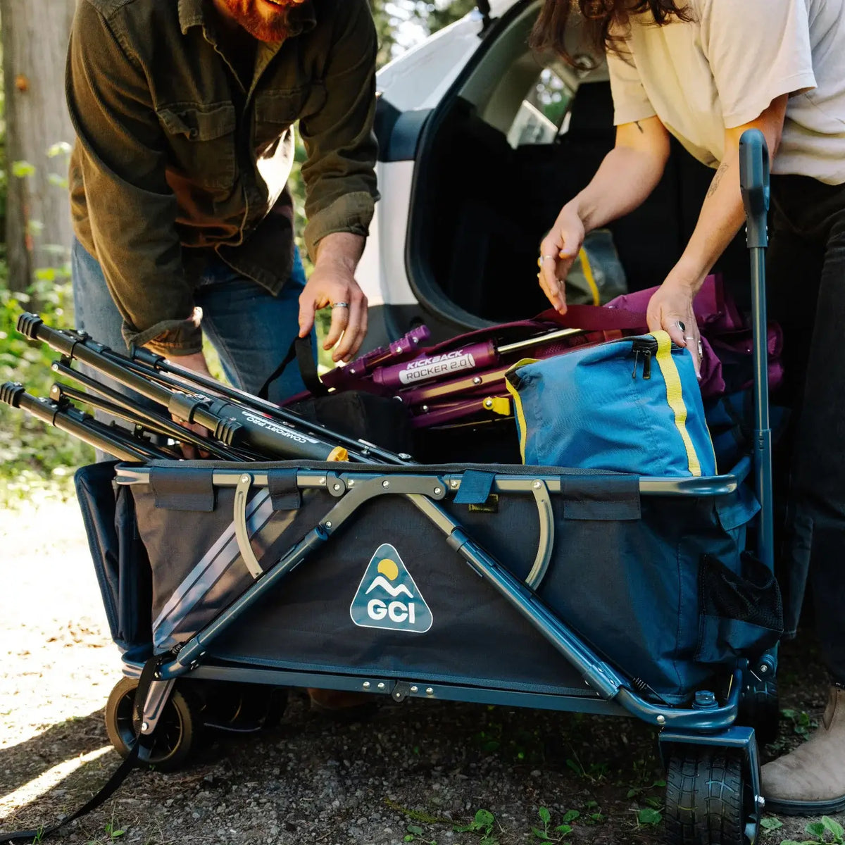 A couple packs gear into an indigo Go-Hauler wagon, showing the folding frame and side pocket in a campsite.