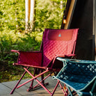 A berry Kickback Insulated Rocker with quilted seating sits on a wooden deck beside a teal chair, near an A-frame cabin in the woods.