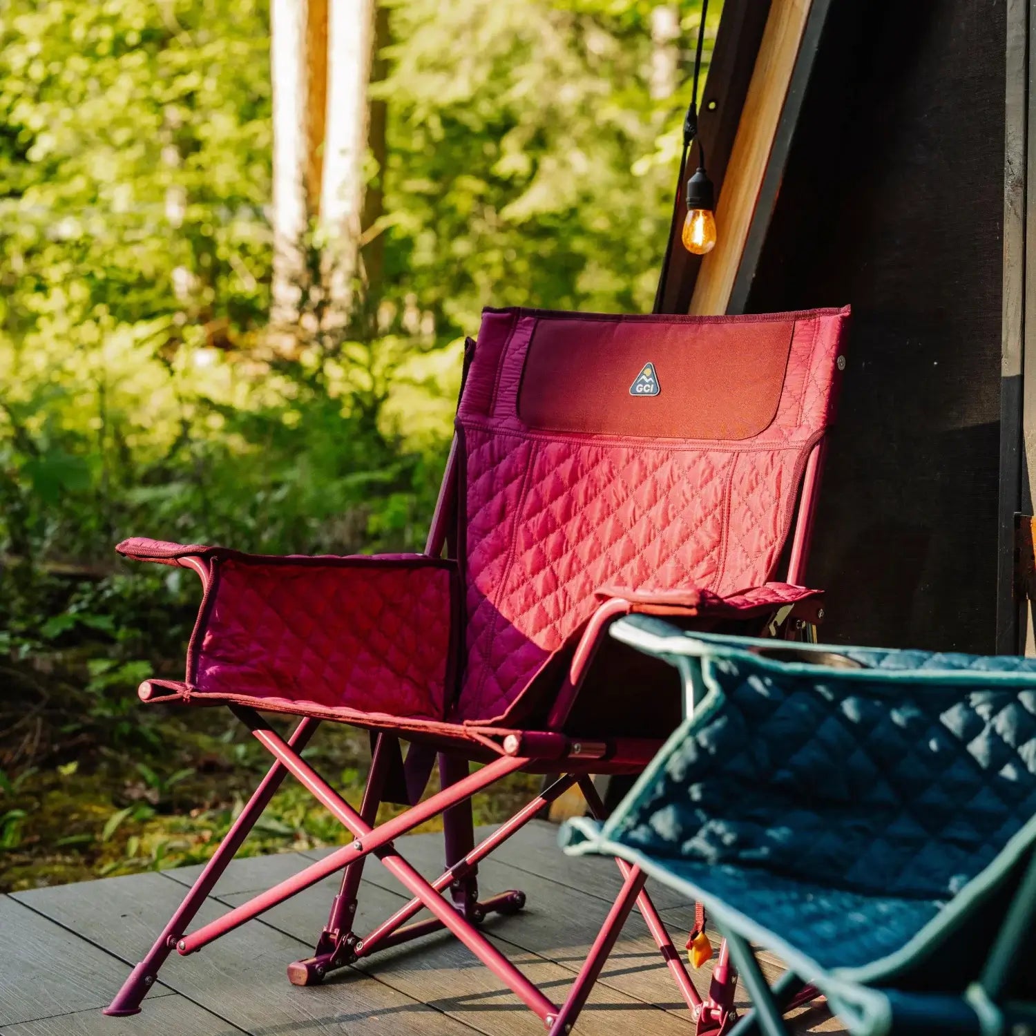 A berry Kickback Insulated Rocker with quilted seating sits on a wooden deck beside a teal chair, near an A-frame cabin in the woods.