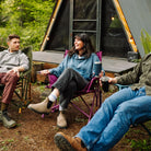 Three friends sit and talk in Kickback Insulated Rockers, including a berry model, on a wooded path near an A-frame cabin, holding metal cups.
